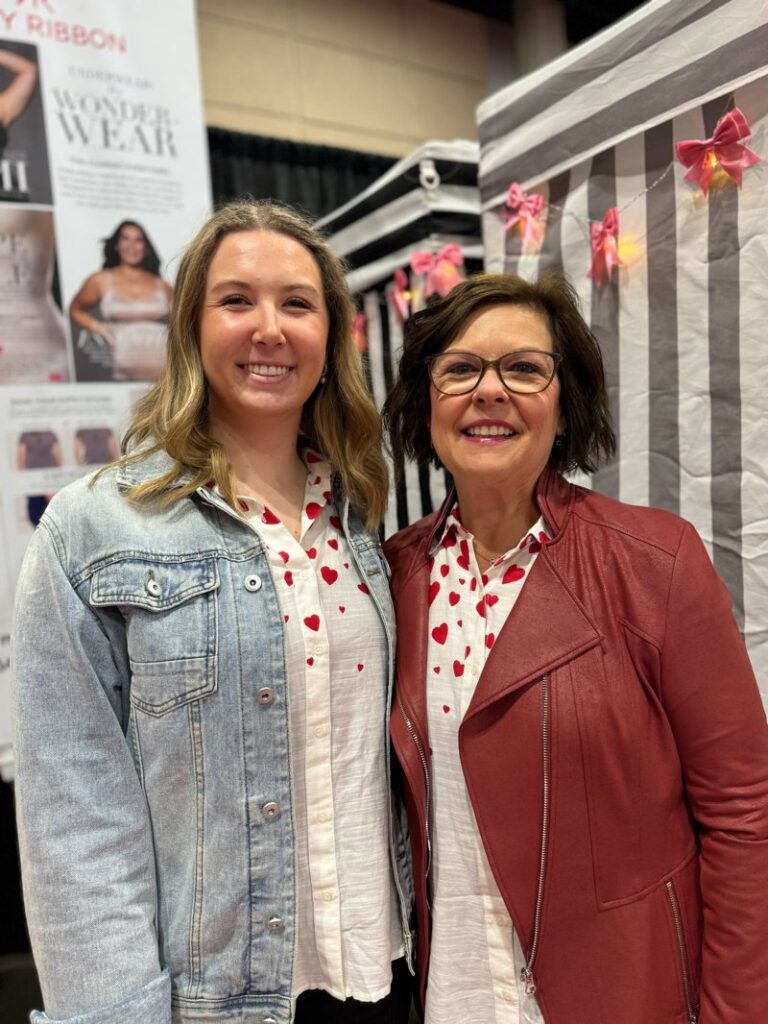 Royanna and her 24-year-old daughter smiling together at a Ruby Ribbon vendor event, both wearing heart-print tops and supporting body confidence for women and a better first bra experience for teens.


